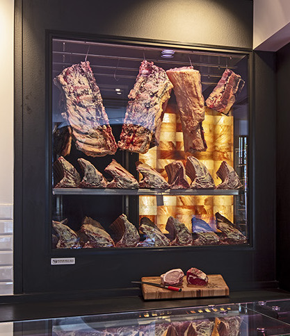 TAR. The Aging Room. Meat Dry-Aging Walk-In Chamber. A wall of Himalayan salt on the back. Meat is hanging and laying on the shelves. Dry-aged steaks are on a cutting board.